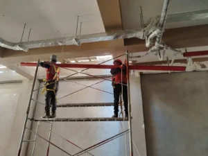 Dos técnicos en andamio instalando tubería contra incendio en un área amplia con ductos