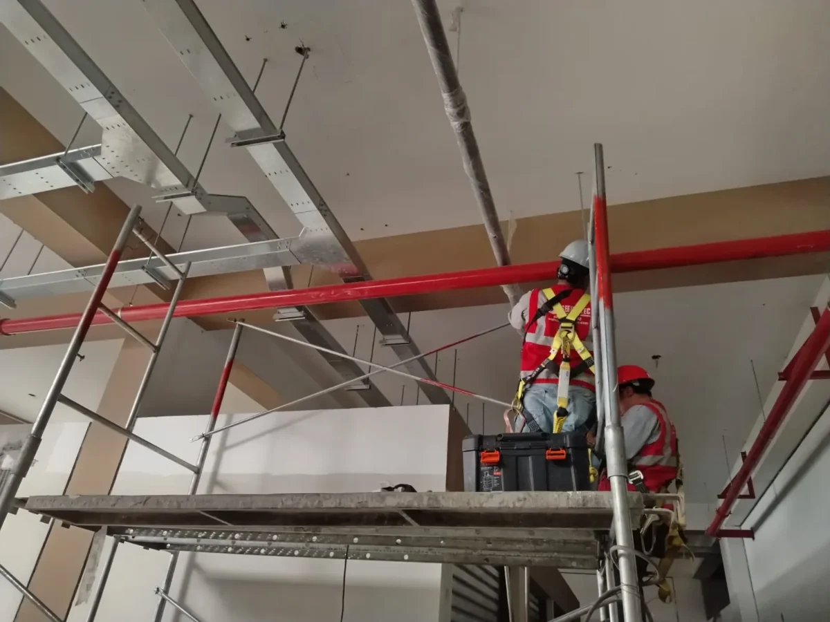 Técnicos en andamio trabajando entre bandejas y tubería roja