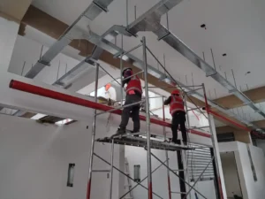 Dos técnicos en andamio colgando tubería contra incendio bajo bandejas y ductos