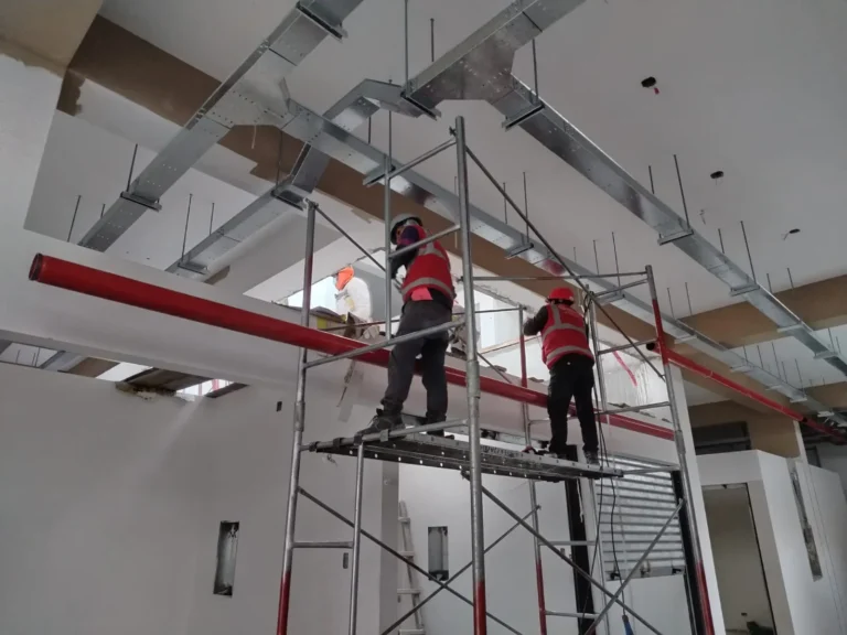 Dos técnicos en andamio colgando tubería contra incendio bajo bandejas y ductos