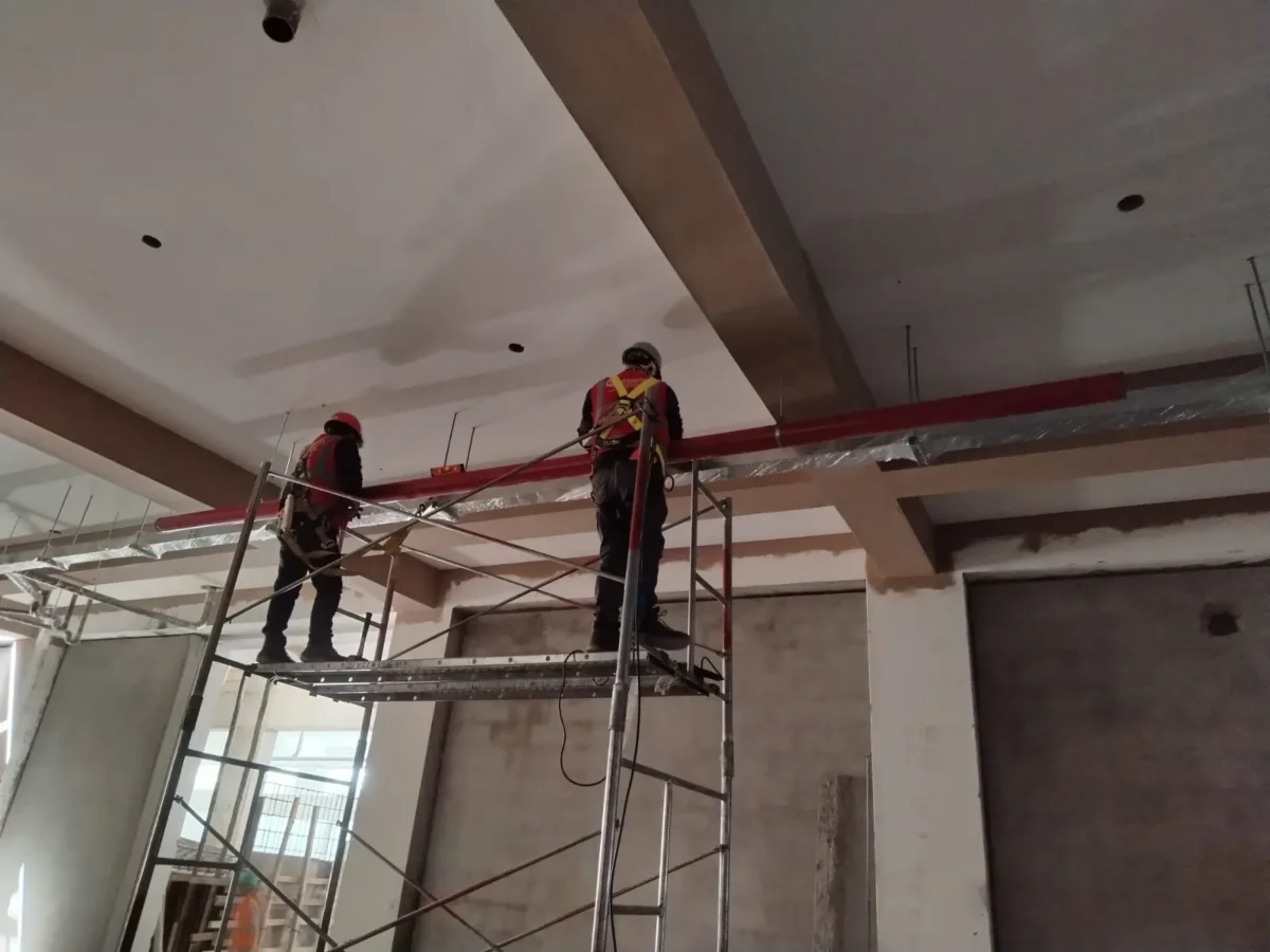 Técnicos sobre andamio instalando tubería roja de agua contra incendio bajo el techo