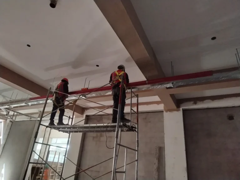 Técnicos sobre andamio instalando tubería roja de agua contra incendio bajo el techo
