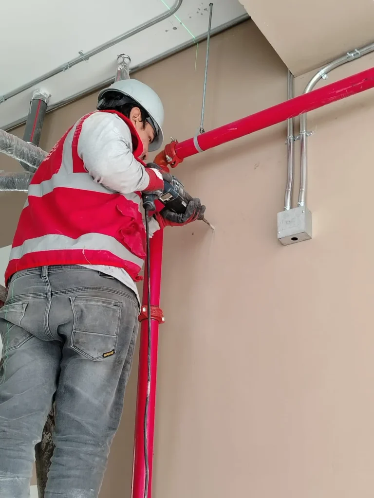 Técnico perforando la pared para el soporte de tubería roja junto a canaletas eléctricas