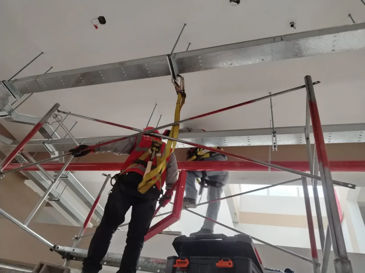 Técnico con arnés en andamio trabajando bajo bandejas metálicas