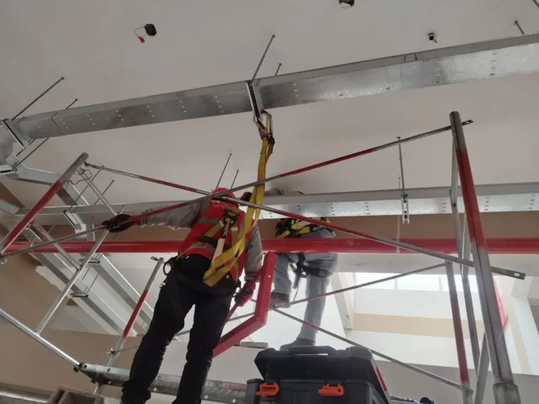 Técnico con arnés en andamio trabajando bajo bandejas metálicas