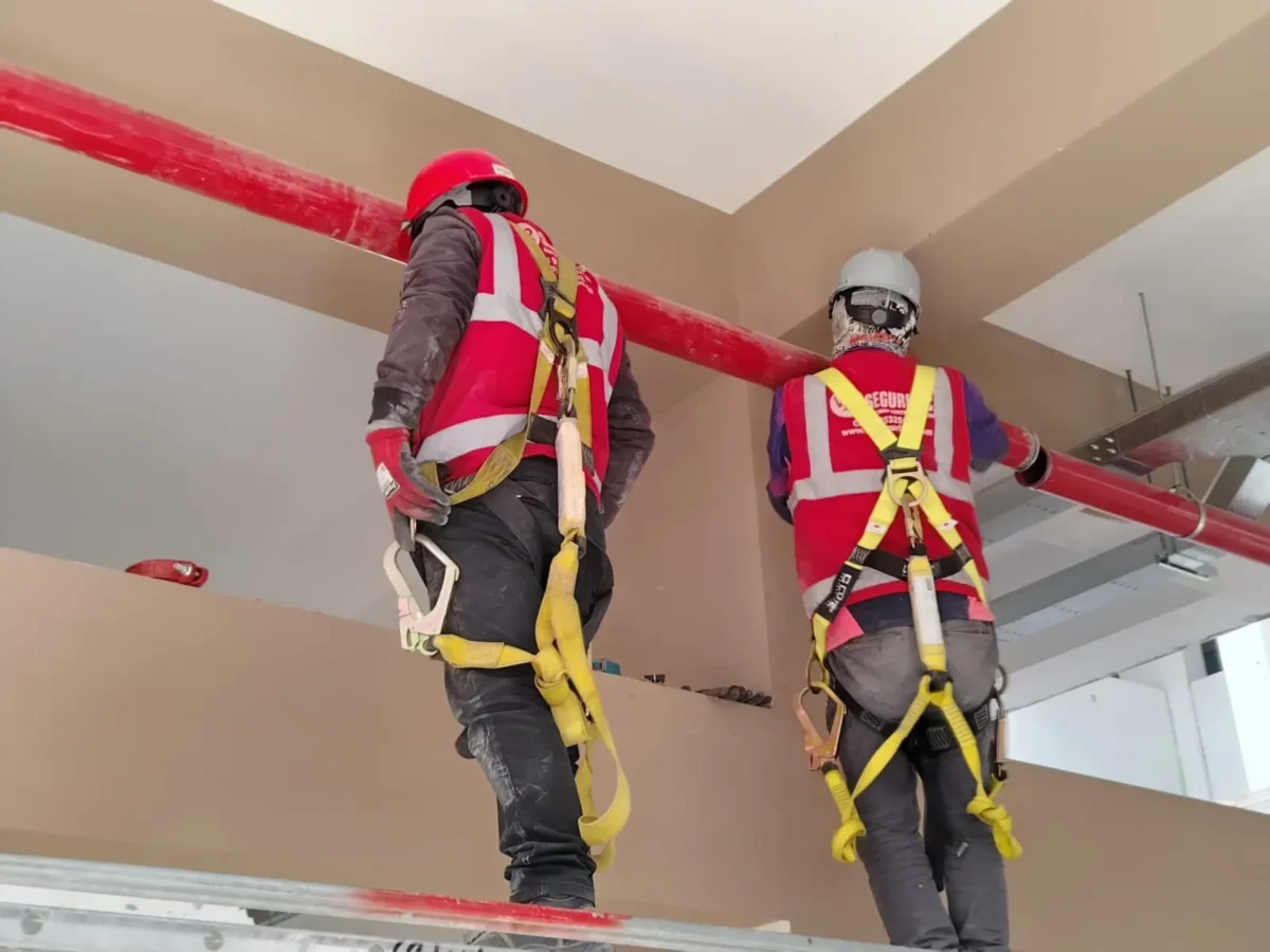Dos técnicos con arnés y casco montando tubería roja sobre una viga