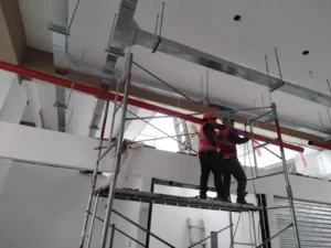 Técnicos con equipo de seguridad montando tubería roja entre ductos de ventilación