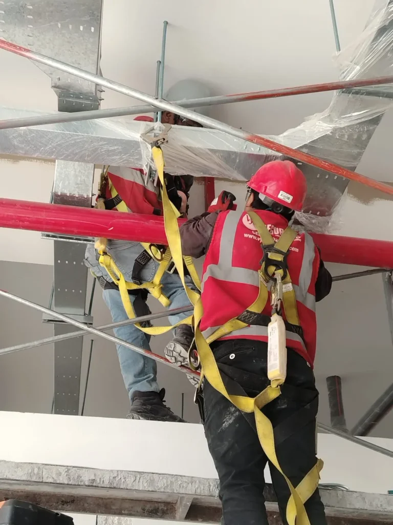 Técnicos con arnés montando tubería roja contra incendio cerca de bandejas portacables galvanizadas
