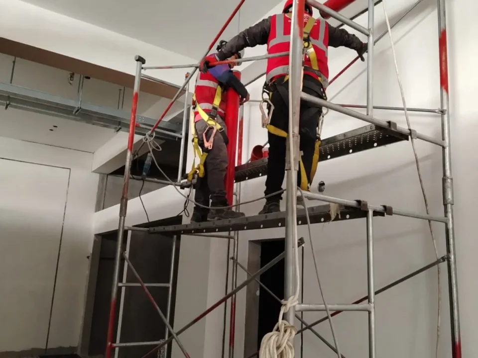 Dos técnicos con arnés sobre un andamio instalando tubería roja de una red contra incendio en un ambiente amplio